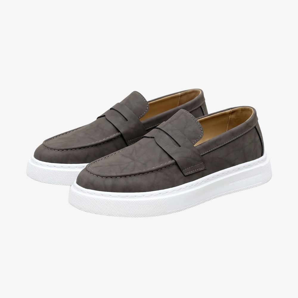 Mocasín de piel marrón para hombre con suela blanca, sobre fondo gris