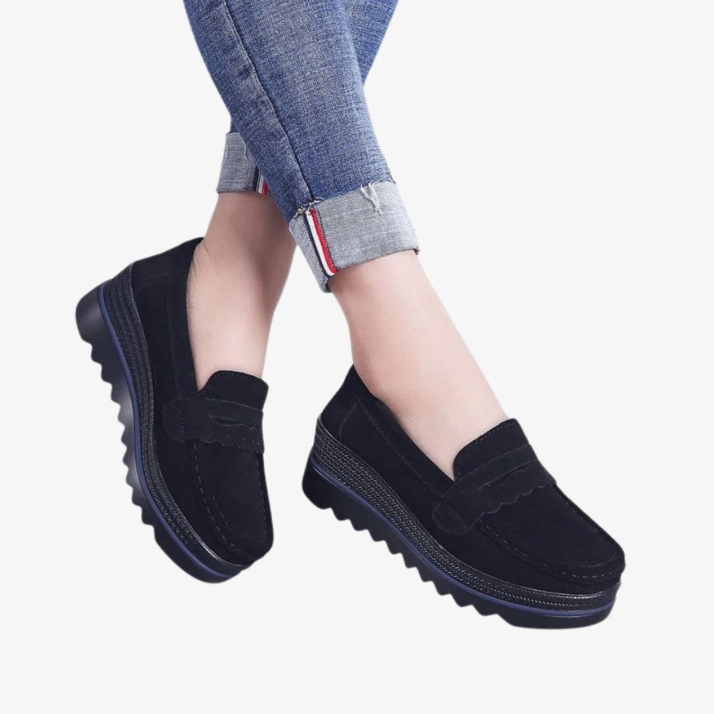Mocasines para mujer en ante negro, elegantes y cómodos, con suelas de cuña con tacos para mayor estabilidad. Su diseño incluye detalles decorativos en la parte superior y una costura visible, perfectos para un estilo moderno e informal.