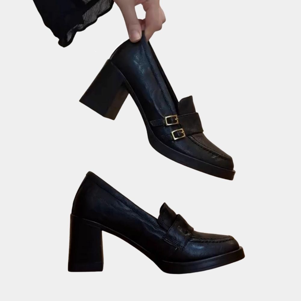 Par de mocasines negros de tacón alto, con un zapato colocado de perfil y el otro sujeto entre los dedos de una mujer