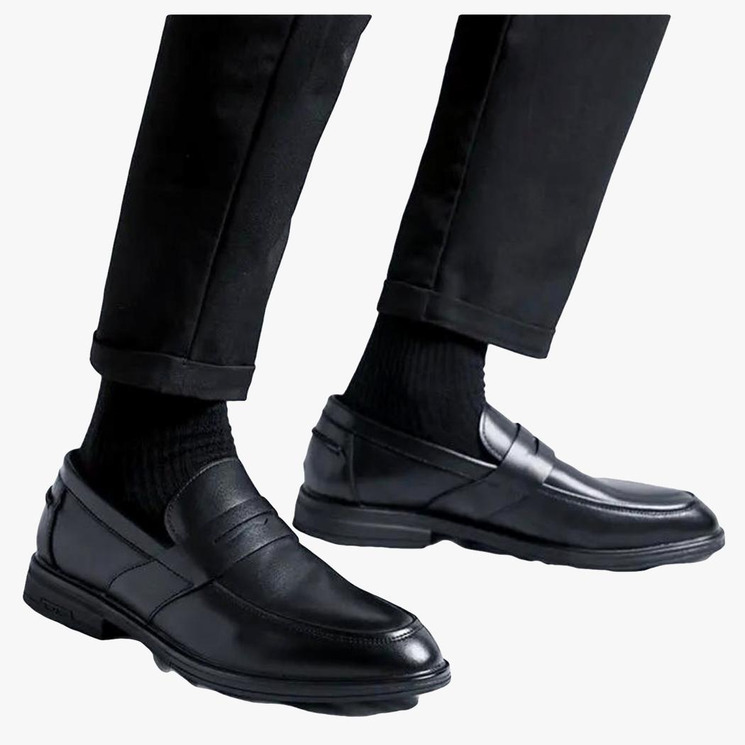 Sobre fondo claro se ve la parte inferior de las piernas de un hombre con pantalón negro y mocasines negros clásicos de piel sintética para hombre.
