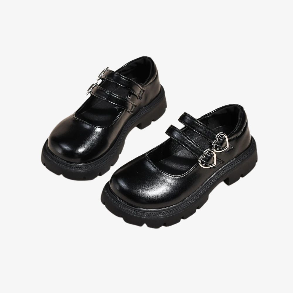 Par de mocasines negros con doble correa y un detalle de corazón encima, sobre fondo gris.