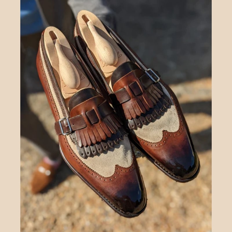 Un par de mocasines semipulidos en piel lisa y ante sintético marrón y beige con flecos bicolores y cierre de correa y hebilla en la parte superior.