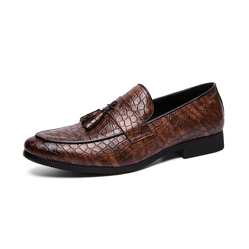 Mocasín de piel de serpiente marrón con una banda en la parte superior y borlas decorativas, sobre fondo blanco.