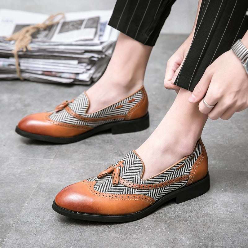 Mocasines bicolores con dibujo de espiga blanco y negro y cuero marrón claro y borlas decorativas en los pies de un hombre con pantalón de traje negro.