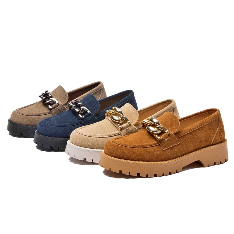 Cuatro mocasines de ante de diferentes colores, gris-azul-beige y marrón, con suelas gruesas y grandes hebillas doradas decorativas alineadas sobre un fondo blanco.