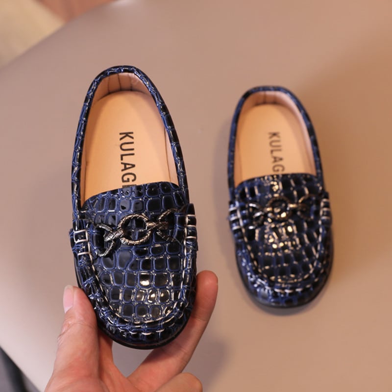 mocasín de bebé en piel sintética con estampado de cocodrilo y una anilla en la parte superior, estos zapatos se presentan en una mano de mujer