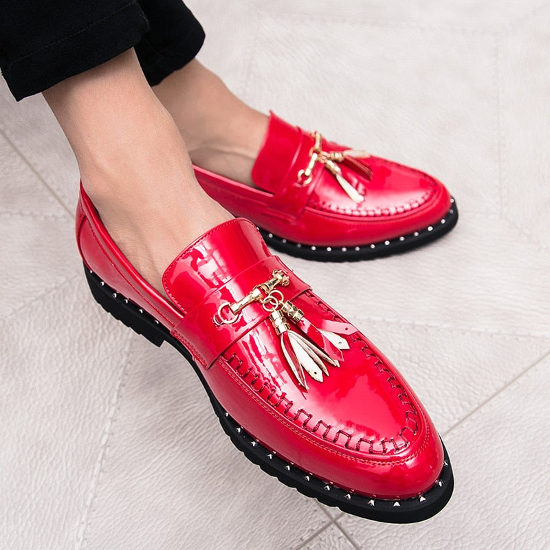 Mocasines de charol rojo brillante con borlas decorativas en los pies de un hombre sentado con las piernas cruzadas que lleva pantalones de traje negros.