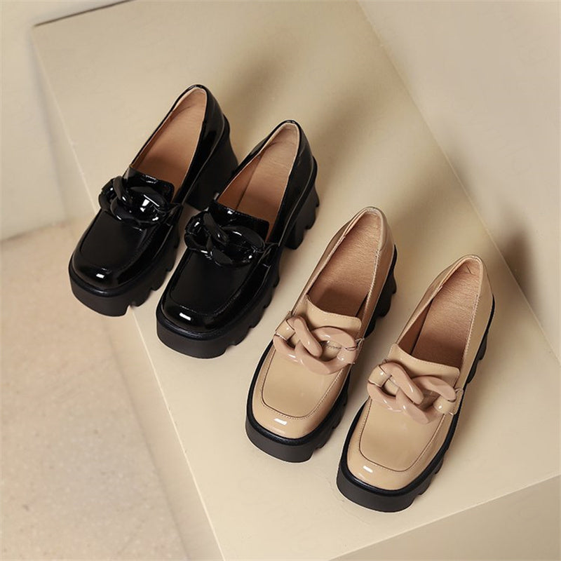 Dos pares de mocasines de charol, uno negro y otro beige, con una hebilla XXL en forma de signo del infinito sobre fondo beige claro.