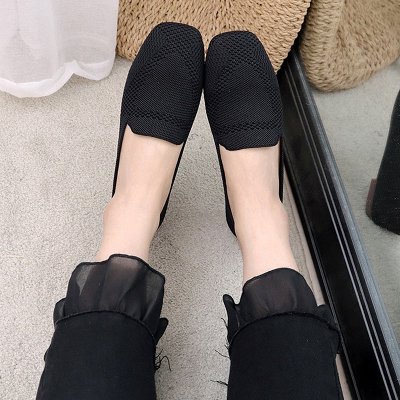 Sobre una mesa de madera, 3 mocasines de punta cuadrada de malla amarilla, negra y beige. Se ve una mano doblando un mocasín para mostrar su flexibilidad