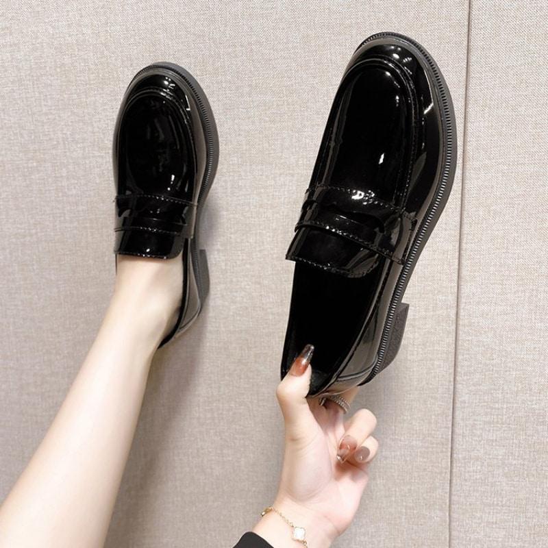Mocasines negros de charol, uno usado y el otro en la mano, sobre fondo gris