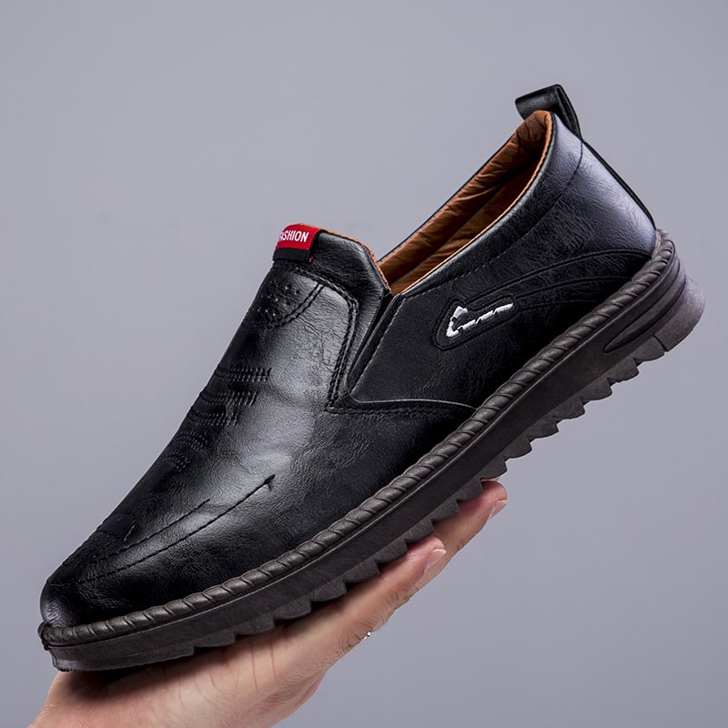 Mocasín negro con suela gruesa, sujeto con una mano. Base gris.