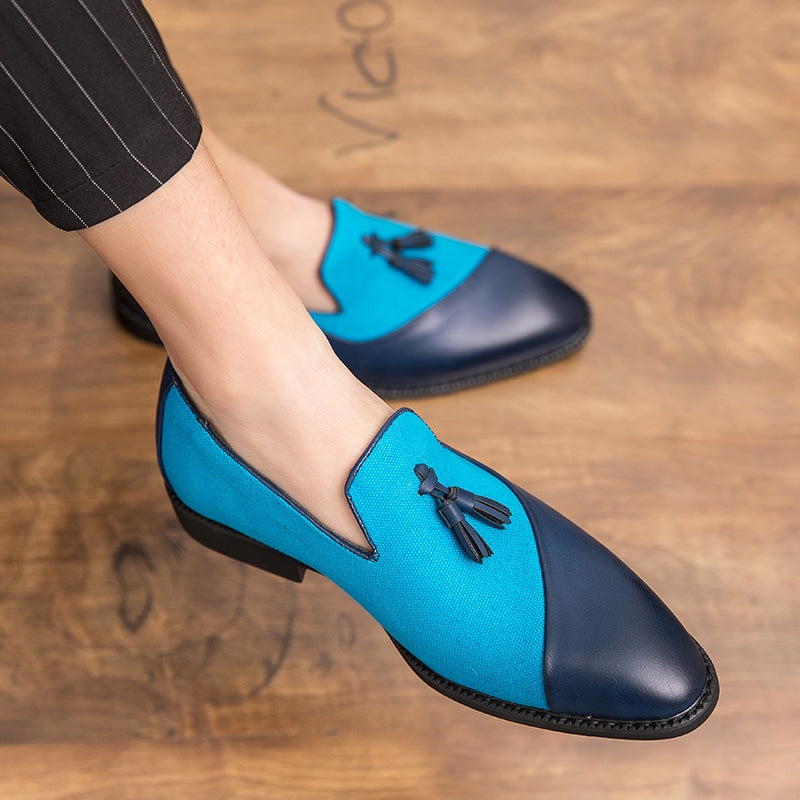 Mocasines de piel y textil azul claro y oscuro en contraste con borlas decorativas en los pies de un hombre con las piernas cruzadas que viste pantalón de traje negro con finas rayas blancas.