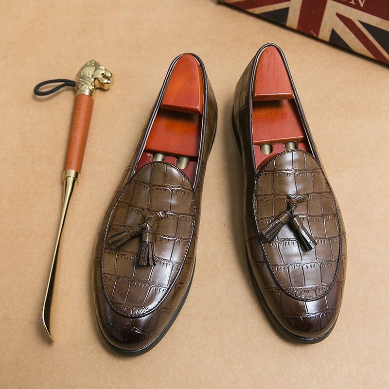 Mocasín de piel de cocodrilo marrón sobre un suelo beige y junto a un calzador