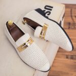 En una caja de madera clara, un par de mocasines blancos alargados con pequeños agujeros y una correa dorada en la parte superior. Dentro de los zapatos hay unas hormas de madera con un botón dorado en el talón.
