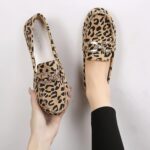 Par de mocasines en estampado de leopardo con pespuntes y anillos de brillantes plateados en la parte superior. Un zapato en un pie y el otro sujeto con una mano.