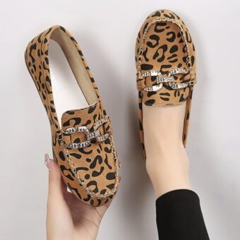 Un par de mocasines redondos con estampado de leopardo marrón y anillos de brillantes plateados, un zapato presentado en el pie con medias negras y el otro sujeto con una mano