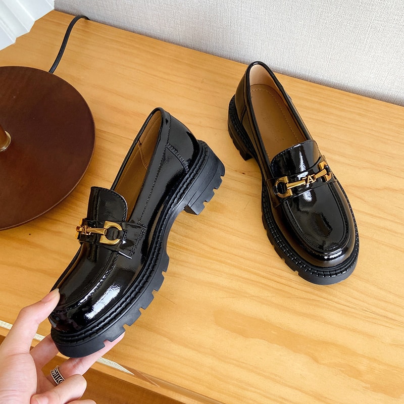 Fotografía de un par de mocasines negros de charol sostenidos por alguien, apoyados en un mueble de madera junto a una lámpara