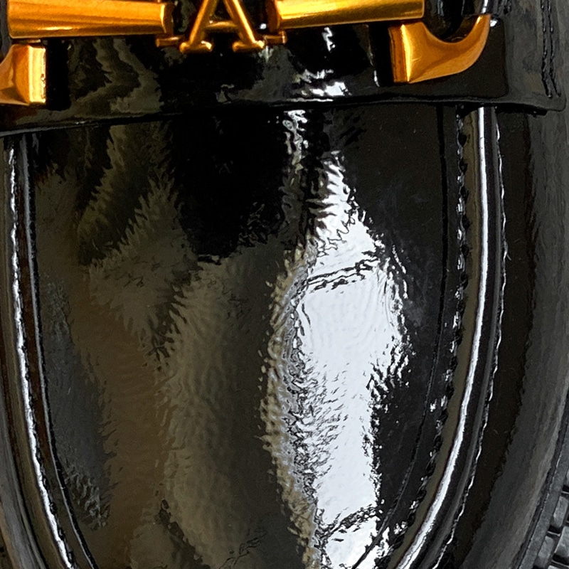 Foto de un zoom sobre un mocasín de charol negro con una cadena dorada y una A