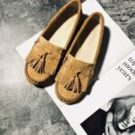 Mocasines de ante camel de estilo indio con flecos y borlas, colocados sobre una revista en un suelo de hormigón pulido.