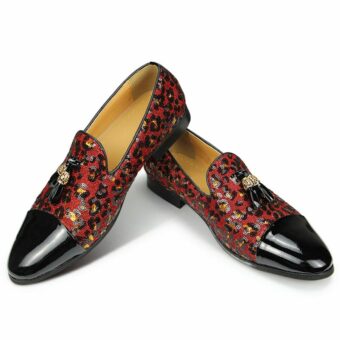 Sobre fondo blanco, un par de mocasines con la parte delantera de charol negro, el resto del zapato de lentejuelas con estampado de leopardo en rojo y borlas en la parte superior. El interior es de color beige