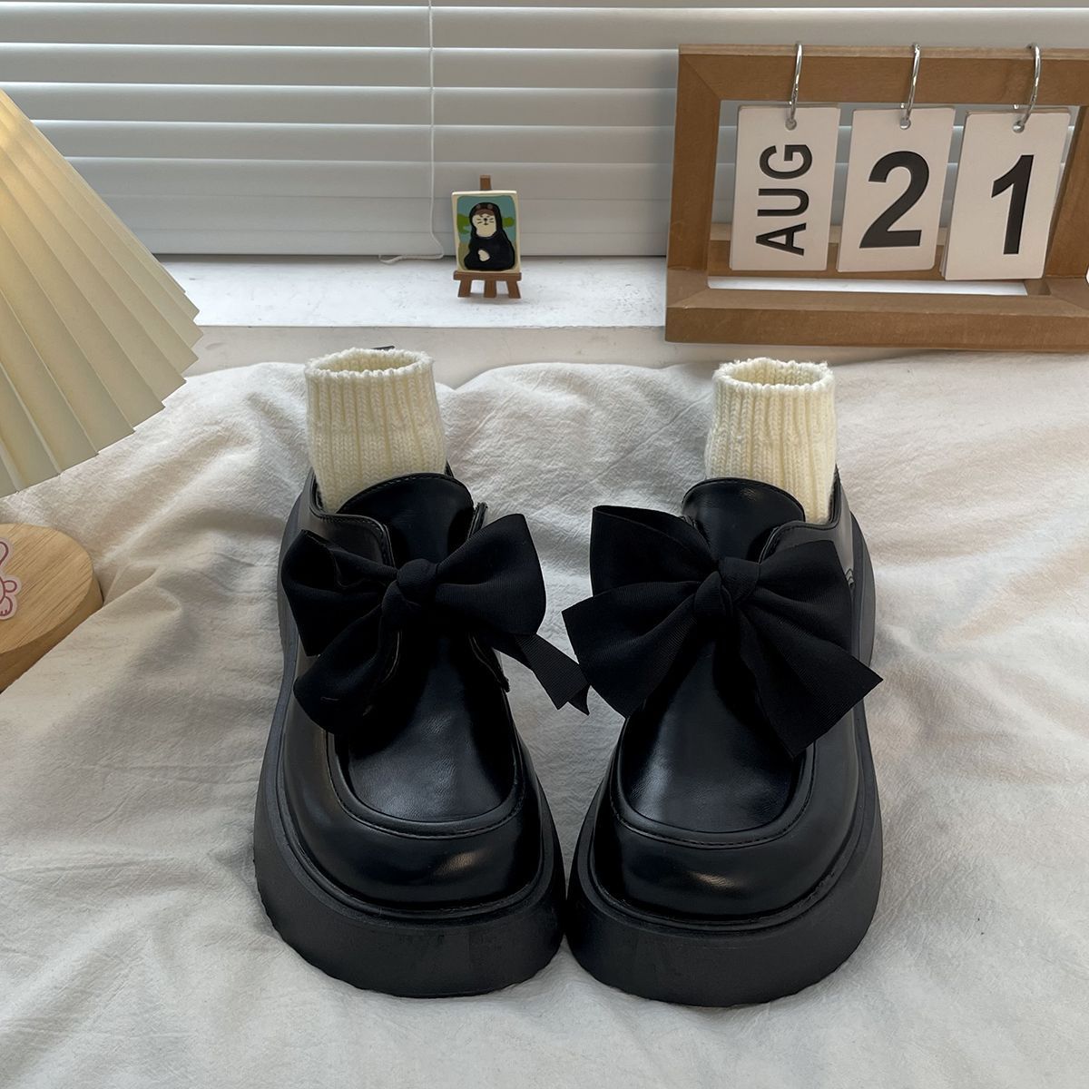 Un par de mocasines de cuero negro liso sobre una cama blanca, con un gran lazo decorando la parte superior y pequeños calcetines beige rellenando el zapato.