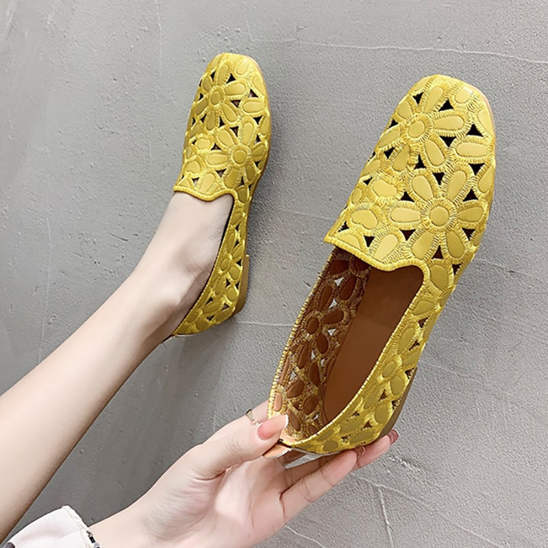Par de mocasines amarillos con motivos florales. Un zapato está en un pie apoyado contra una pared gris y el otro está sujeto con una mano