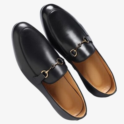 Mocasín negro con hebilla dorada en piel vegana sobre fondo blanco roto.